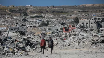 Two people walk through a vast landscape of destruction and rubble, with collapsed buildings around them.