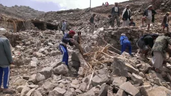 L'image montre un groupe de personnes travaillant dans les décombres d'un bâtiment effondré. Elles semblent s'affairer à dégager des pierres et des débris, avec des outils improvisés. L'environnement paraît montagneux et accidenté, avec des restes de structures anciennes en arrière-plan. L'atmosphère est celle de l'effort collectif dans un contexte de reconstruction ou d'aide humanitaire.