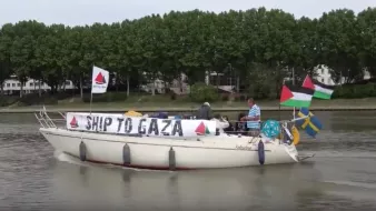 The image shows a boat on a body of water with several individuals on board. The boat has a prominent banner that reads "SHIP TO GAZA," and it is adorned with various flags, including the Palestinian flag and the flag of Sweden. In the background, there are trees and buildings along the shoreline. The scene conveys themes of solidarity and activism related to Gaza.