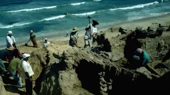 A group of people excavates a sandy site near the ocean, surrounded by waves and tools.