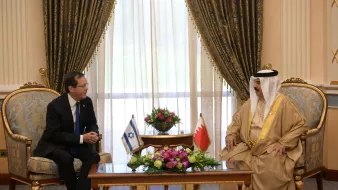 Two men in formal attire sit across from each other in a richly decorated room, discussing.