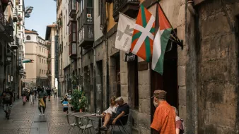 Calle estrecha con gente, mesas al aire libre y banderas ondeando en un ambiente urbano.