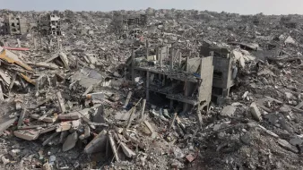 L'image montre un vaste paysage de débris et de bâtiments effondrés, engloutis dans les gravats. On aperçoit des structures en béton brisées et une atmosphère de destruction désolante.