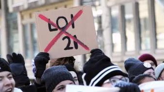 La imagen muestra a un grupo de personas participando en una manifestación. Algunas de ellas sostienen carteles, y uno de los más destacados es un cartel de cartón que dice "X" sobre el número "21", lo que indica oposición a esa ley o número específico. La multitud parece estar animada y comprometida con la causa que defienden. Las personas están abrigadas, usando gorros y guantes, lo que sugiere que hace frío.