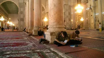 L'image montre l'intérieur d'une grande mosquée, caractérisée par de hauts plafonds et de majestueuses colonnes. Le sol est recouvert de tapis, et plusieurs personnes sont assises sur le sol, certaines concentrées sur leurs ordinateurs portables, tandis que d'autres semblent prier ou méditer. La lumière naturelle entre par les fenêtres, créant une ambiance calme et sereine. Des lustres pendent du plafond, ajoutant une touche d'élégance à cet espace sacré.