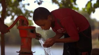 The image shows a young boy bent over a red hand pump, drawing water from it. He appears focused as he cups his hands to catch the water flowing out. The background features greenery and suggests a natural setting, possibly near a body of water, with soft light creating a warm atmosphere. The boy is wearing a red shirt, and the scene conveys a sense of simplicity and the importance of access to fresh water.