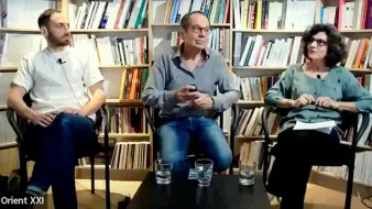 Cette image montre trois personnes assises sur des chaises dans un environnement intérieur, probablement une librairie ou un espace de discussion. À l'arrière-plan, il y a des étagères remplies de livres. La personne à gauche porte une chemise blanche, celle du centre est en chemise grise et celle à droite porte un haut vert. Ils semblent être en train de discuter ou de débattre, avec des verres d'eau devant eux. L'ambiance semble détendue et propice à l'échange d'idées.