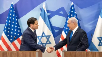 The image shows two men shaking hands at a press conference. They are standing at podiums, with Israeli and American flags in the background.