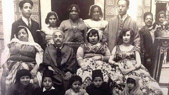 L'image présente un groupe de personnes, probablement une famille ou une communauté, posant ensemble dans un décor intérieur. On peut voir des hommes et des femmes de différentes générations, ainsi que des enfants, vêtus de tenues traditionnelles. L'atmosphère semble conviviale et chaleureuse. L'arrière-plan est orné de motifs, ajoutant une touche historique à la scène.