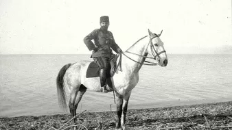 L'image montre un homme en uniforme militaire monté sur un cheval blanc. Il se tient debout sur la plage, près d'une étendue d'eau. L'homme porte un couvre-chef distinctif, et son uniforme semble traditionnel. Le paysage est simple, avec des galets au premier plan et un horizon calme en arrière-plan. L'atmosphère donne une impression de sérénité et de majesté.