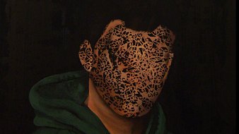 L'image montre un portrait d'une personne avec un visage obscurci par un motif complexe en noir et doré. La personne porte un sweat à capuche vert. L'arrière-plan est sombre, ce qui met en évidence le contraste entre le visage décoratif et le reste de l'image. L'aspect du visage semble à la fois mystérieux et artistique, créant une impression intrigante.