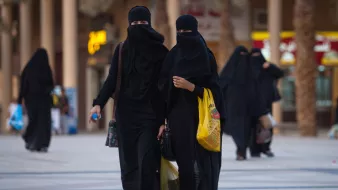 L'image montre deux femmes marchant côte à côte dans une zone urbaine. Elles portent des vêtements noirs, y compris des niqabs qui couvrent leur visage, laissant seulement les yeux visibles. L'environnement semble animé, avec des bâtiments en arrière-plan et d'autres personnes se déplaçant dans la même direction. L'une des femmes tient un sac, tandis que l'autre semble tenir un objet dans sa main. L'éclairage et l'architecture suggèrent un cadre contemporain.