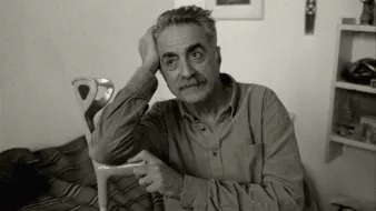 Homme pensif, assis, avec une canne, dans une pièce simple et lumineuse.
