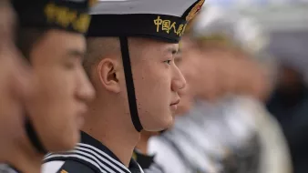 L'image montre des marins en uniforme, probablement lors d'une cérémonie ou d'un événement officiel. On peut voir des hommes alignés, portant des chapeaux distinctifs et des uniformes marins. Les expressions sur leurs visages sont sérieuses, et ils semblent concentrés sur un point donné. L'arrière-plan montre des silhouettes floues d'autres marins, suggérant un grand rassemblement. L'image dégage une ambiance de discipline et de camaraderie militaire.