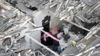 L'image montre des ruines d'un bâtiment, avec des débris de toit et des murs effondrés. Deux personnes sont visibles à l'intérieur des décombres, semblant chercher ou s'entraider dans cet environnement chaotique.