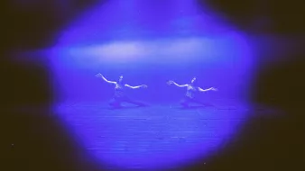 The image features two dancers positioned on a stage, surrounded by a soft, dramatic purple light. They appear to be performing a synchronized dance, with their arms outstretched. The background is dark, drawing focus to the illuminated area where they are dancing. The scene conveys a sense of artistry and expression, emphasizing the dancers' movements and the emotional atmosphere created by the lighting.