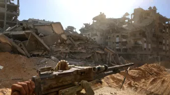 تصویری از یک منطقه ویرانشده، با ساختمانهای خراب و تفنگی در پیشزمینه. غبار و تابش نور خورشید در آسمان.