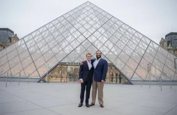 L'image montre deux hommes posant ensemble devant le Musée du Louvre, qui est célèbre pour sa pyramide en verre. Le bâtiment est moderne et impressionnant, contrastant avec l'architecture classique des ailes du musée. Les hommes sourient et portent des costumes formels, se tenant côte à côte, tandis que le ciel est légèrement nuageux, ajoutant une atmosphère calme à la scène.