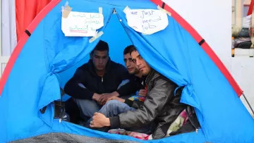 L'image montre un groupe de trois jeunes hommes assis à l'intérieur d'une tente. Ils semblent discuter ensemble, et des pancartes en arabe et en anglais sont visibles à l'extérieur de la tente, indiquant "Nous avons juste besoin d'un emploi". La tente est de couleur bleue, et l'ambiance générale évoque un sentiment de rassemblement autour d'une cause.