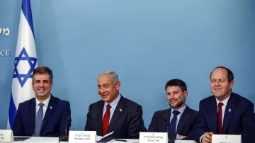 The image shows a group of four men seated at a table in front of a blue backdrop with the Israeli flag. They appear to be engaged in a discussion or announcement. The man in the center, likely the most prominent figure, is smiling. Each individual has a nameplate in front of them, indicating their identities or titles. The overall atmosphere seems formal, suggesting a political or governmental context.