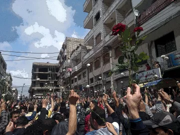 L'image montre une grande foule au milieu d'une rue, probablement lors d'une manifestation. Les gens semblent levés les bras, tenant des roses, ce qui suggère une ambiance pacifique ou festive. En arrière-plan, on aperçoit des bâtiments et un ciel partiellement nuageux. L'atmosphère semble être chargée d'émotions et d'unité entre les participants.