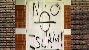 L'image montre une porte sur laquelle est peinte une inscription en spray noir. Le mot "NO" est écrit en haut à gauche, suivi d'un symbole ressemblant à une croix, et en dessous, le mot "ISLAM" est inscrit avec un point d'exclamation à la fin. Le fond de la porte et le mur environnant montrent des briques. L'ensemble de la scène évoque un message provocateur.