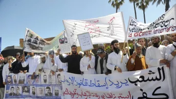 L'image montre une manifestation avec un groupe de personnes tenant des pancartes et des banderoles. Les manifestants semblent exprimer des revendications politiques ou sociales, avec des slogans écrits en arabe et en français. On peut voir des visages déterminés et des gestes de protestation. La scène est entourée d'arbres, suggérant un environnement urbain. L'atmosphère est chargée d'émotion, reflétant l'engagement des participants.