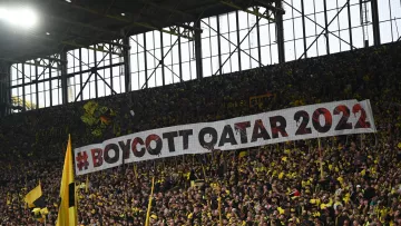 L'immagine mostra una folla di tifosi all'interno di uno stadio, con un grande striscione che recita "#BOYCOTT QATAR 2022". La maggior parte dei tifosi indossa magliette gialle e nere, colori tipici della loro squadra. L'atmosfera sembra essere intensa e carica di emozione, con un forte senso di protesta evidenziato dallo striscione.