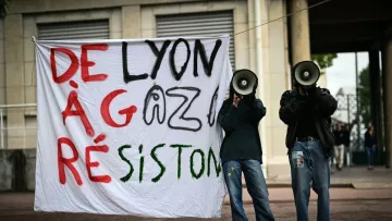 L'image montre une manifestation avec une grande pancarte accrochée qui dit "DE LYON À GAZA RÉSISTONS". Deux personnes se tiennent derrière la pancarte, chacune tenant un mégaphone. L'environnement semble urbain, et l'atmosphère suggère un engagement pour une cause.