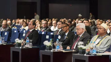 L'image montre une assemblée de personnes assises dans une salle de conférence. Les participants semblent attentifs et engagés, avec des tables devant eux, ornées de petites fleurs et de bouteilles d'eau. On peut voir plusieurs hommes en costume, ainsi que des femmes portant des vêtements formels. L'environnement est élégant, avec des chaises bleues et une disposition ordonnée. Les visages des participants expriment une concentration sur l'événement en cours.