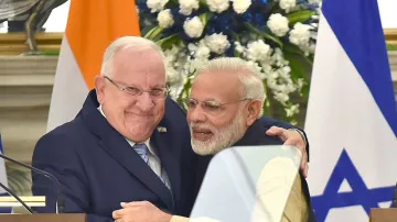 L'image montre deux hommes en train de se serrer dans les bras, exprimant une atmosphère chaleureuse et amicale. L'un des hommes porte un costume sombre avec une cravate, tandis que l'autre est vêtu d'une tenue traditionnelle indienne. En arrière-plan, on peut voir des drapeaux de l'Inde et d'Israël ainsi que des fleurs décoratives, ce qui indique un contexte officiel ou diplomatique.