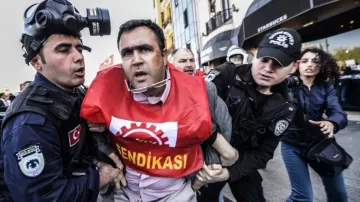 L'image montre une scène de répression où des policiers en tenue d'intervention arrêtent un homme qui porte un t-shirt rouge avec le mot "SENDIKASI". L'individu semble être en train d'être mené à l'écart, visiblement en désaccord avec son arrestation. Les policiers sont équipés de casques et semblent concentrés sur leur opération. L'environnement est urbain, et on peut voir un café Starbucks en arrière-plan, ce qui suggère que cette scène se déroule dans une zone animée de la ville.