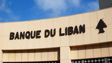 L'image montre un bâtiment avec l'inscription "BANQUE DU LIBAN" en lettres majuscules. La façade est faite de pierre ou de béton, et il y a un symbole d'un petit arbre à côté du texte. Le ciel en arrière-plan est bleu avec quelques nuages. L'ensemble donne une impression de modernité et de solidité.