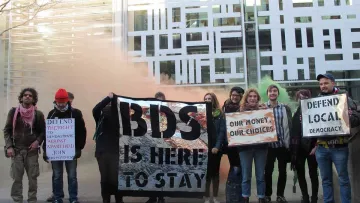 The image depicts a group of protesters standing outside a modern building, holding various signs. The signs convey messages related to support for the BDS movement, defending local democracy, and advocating for financial choices. The atmosphere seems charged, possibly with smoke or color in the background, emphasizing the protest's fervor. The group consists of diverse individuals, some wearing casual attire, and they appear to be unified in their cause.