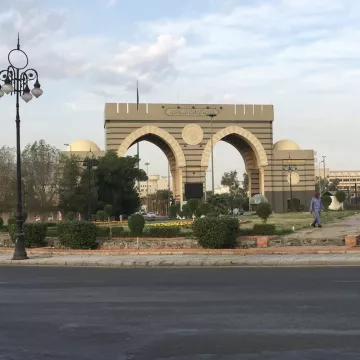 Un grand portail avec arches, entouré de verdure et de lampadaires, sous un ciel nuageux.
