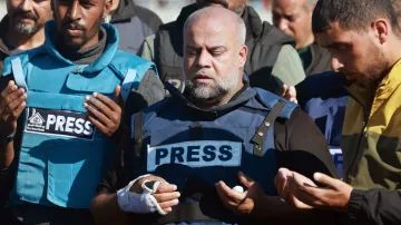 The image depicts a group of individuals, including a central figure wearing a blue vest marked "PRESS." This person appears to be in a moment of prayer or contemplation, with a serious expression, and has a bandaged hand. Surrounding him are others also dressed in similar press attire, some raising their hands in a gesture of prayer. The setting suggests a somber or reflective atmosphere, likely related to a significant event or situation involving journalists.