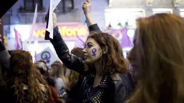 L'image montre un groupe de personnes participant à une manifestation. Au premier plan, une jeune femme avec des cheveux bouclés et un maquillage marqué, porte une veste en cuir et tient un morceau de papier ou une affiche. Son visage est décoré d'un symbole féminin en violet, qui se retrouve également sur d'autres bannières en arrière-plan. L'atmosphère semble être celle d'un rassemblement pour les droits des femmes, soulignant la solidarité et l'engagement des participants.