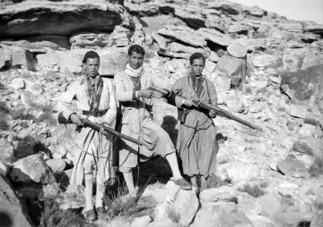 L'image montre trois hommes vêtus de vêtements traditionnels, tenant chacun un fusil. Ils se tiennent debout sur un sol rocheux, entourés de grandes pierres et de formations rocheuses. L'environnement semble austère et désertique. Les hommes ont des expressions sérieuses et sont alignés côte à côte. Leur posture et l'équipement qu'ils portent suggèrent qu'ils peuvent être des membres d'un groupe armé ou des guerriers appartenant à une communauté spécifique.