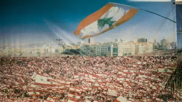 L'image montre une grande foule rassemblée, probablement lors d'une manifestation ou d'un événement public au Liban. La mer de personnes brandit de nombreux drapeaux libanais, créant un effet visuel saisissant. En arrière-plan, on aperçoit une ville moderne, ce qui suggère une ambiance vibrante. Le ciel est dégagé, ajoutant à la luminosité de la scène. Cette image évoque un sentiment de patriotisme et de solidarité parmi les participants.