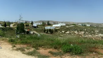 L'image montre un paysage rural avec des collines à l'arrière-plan. On peut voir plusieurs maisons en métal ou modulaires, disposées de manière dispersée. La végétation est sparse, avec des zones de terre et de l'herbe verte. Le ciel est clair et ensoleillé. L'ensemble donne une impression d'isolement et de simplicité.