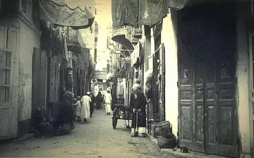 L'image montre une ruelle étroite d'une ville ancienne, probablement dans un pays arabe. Les murs des bâtiments sont ornés de tentures et de draps qui ajoutent une ambiance colorée. On peut voir des habitants, dont certains portent des vêtements traditionnels, déambulant dans la rue. Un homme semble se tenir près d'une charrette, tandis que d'autres personnes se déplacent en arrière-plan. Les portes en bois, typiques, ajoutent un charme ancien à la scène, qui évoque une atmosphère de vie quotidienne dans un vieux quartier.