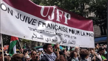 L'image montre une manifestation où des personnes tiennent une grande banderole. Cette banderole mentionne "UJFP" et affiche des messages en français, hébreu et arabe, qui semblent appeler à la paix. Le contexte suggère un rassemblement pour des causes sociales ou politiques, probablement en lien avec la paix au Moyen-Orient. En arrière-plan, on voit une foule de manifestants, des arbres et des bâtiments, ce qui indique que l'événement se déroule en milieu urbain.