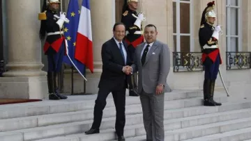 L'image montre une scène officielle à l'extérieur d'un bâtiment gouvernemental. Deux hommes, en costume, se serrent la main sur des marches, symbolisant une rencontre diplomatique. À l'arrière-plan, on peut voir des drapeaux de la France et du Maroc, ainsi que des soldats en tenue d'apparat. L'atmosphère est formelle et solennelle, reflétant l'importance de l'événement.