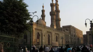 L'image montre un vaste bâtiment architectural au style islamique, probablement une mosquée, avec des minarets majestueux s'élevant vers le ciel. On peut voir des personnes en tenue traditionnelle, certaines portant le hijab. En arrière-plan, la lumière du soleil est douce, créant une atmosphère paisible. Les détails architecturaux de la façade en pierre ajoutent une touche historique à la scène. Des arbres et des lampadaires embellissent également l'environnement.