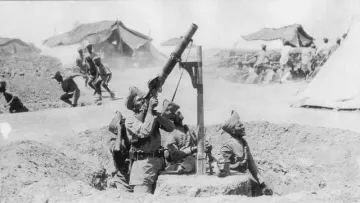 L'image en noir et blanc montre un groupe de soldats en position de tir. Un soldat est en train d'opérer un mortier, levé vers le ciel, tandis que deux autres soldats se trouvent à proximité, probablement prêts à aider ou à surveiller la situation. En arrière-plan, on peut voir des tentes militaires, indiquant qu'ils sont dans un camp. D'autres soldats semblent en mouvement, suggérant une activité intense ou une opération en cours. L'atmosphère générale de l'image évoque un contexte militaire et historique.