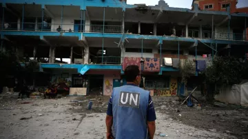 The image depicts a scene of destruction, likely from a conflict area. In the foreground, a person wearing a blue vest with "UN" printed on it stands, facing a damaged building with shattered windows and debris scattered around. The building has signs of neglect, with graffiti and remnants of what may have been a children's play area. People can be seen in the background, possibly assessing the situation or providing assistance. The overall atmosphere conveys a sense of urgency and the aftermath of turmoil.