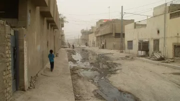 L'image montre une rue étroite et déserte dans un quartier urbain, probablement en milieu rural. Les bâtiments qui l'entourent semblent en mauvais état, avec des murs en béton et des façades peu entretenues. Le sol est inégal et présente des flaques d'eau, ce qui indique probablement une certaine négligence ou des problèmes d'infrastructure. L'atmosphère est assez grise et brumeuse, suggérant une faible visibilité, possiblement à cause de la poussière ou de la pollution. Un enfant est visible au bout de la rue, marchant seul.