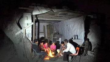 L'image montre un groupe de personnes assises autour d'un feu dans un environnement sombre. Ils semblent rassemblés à l'intérieur d'une structure rudimentaire, faite de matériaux simples comme du bois et des tôles. Les gens, probablement des adultes et des enfants, sont concentrés sur le feu, qui éclaire leur visage et crée une atmosphère chaleureuse malgré les conditions environnantes. Des éléments comme des bouteilles sont visibles en arrière-plan, suggérant une ambiance de partage et de convivialité, malgré le manque de confort.