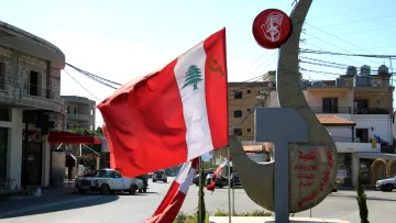 La imagen muestra una intersección en una localidad, con una bandera de Líbano ondeando en el viento. A la derecha, hay una estructura que parece una escultura o un monumento, posiblemente con inscripciones en árabe. Al fondo se pueden ver edificios y postes de luz, mientras que el cielo es despejado y soleado. La escena refleja un ambiente urbano característico de la región.