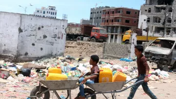 The image shows two boys in a dilapidated urban setting. One boy is pushing a wheelbarrow that carries another boy sitting inside it. The background includes a scene of garbage and rubble, with partially constructed buildings and damaged structures. The environment appears to be neglected, highlighting issues related to poverty and urban decay. The sky is clear, suggesting a bright day.